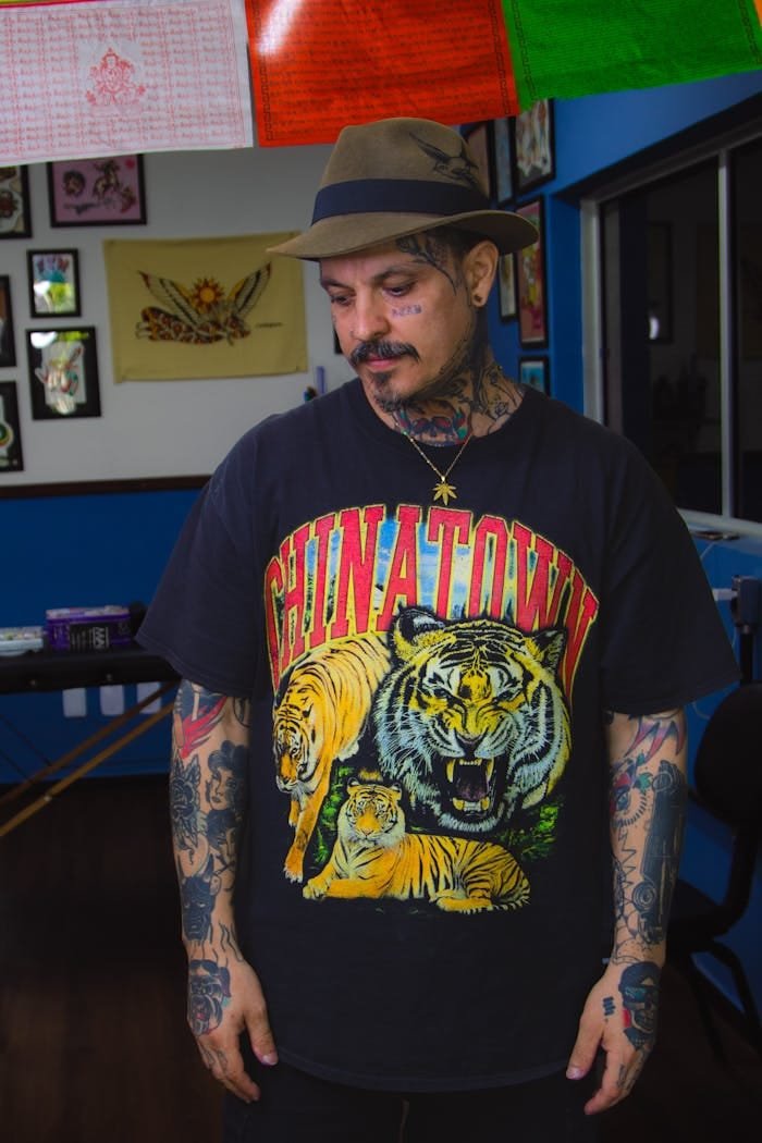 Tattooed man wearing a Chinatown t-shirt with tiger print, standing indoors with hat and mustache.